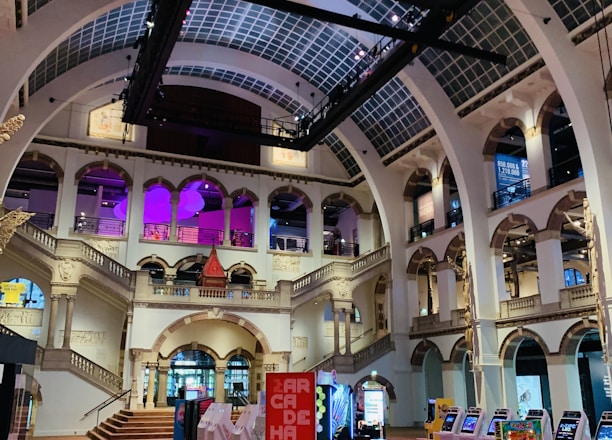 The interior of a large, ornately designed building with arched ceilings and intricate architectural details. The space features gaming machines lined up along the floor, under a high ceiling with a grid pattern. Lighting in purple and white tones enhances the ambiance.