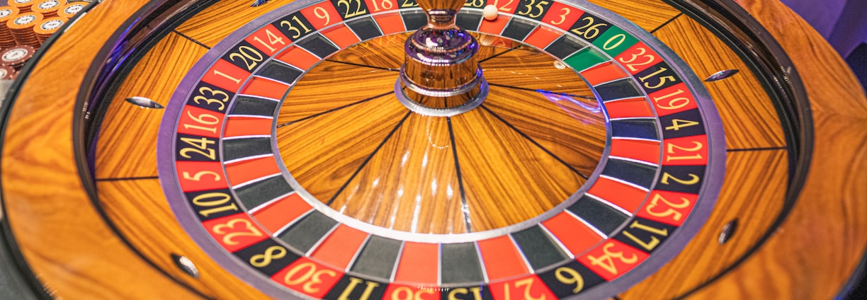 A roulette wheel with alternating red and black numbered pockets on a wooden platform. The wheel contains a small white ball resting in one of the pockets, indicating gameplay. Surrounding the wheel are stacks of colorful poker chips, suggesting a casino environment.