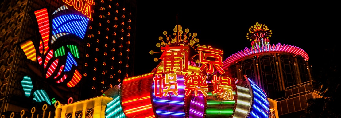 Bright neon lights and colorful signage adorn the exterior of Casino Lisboa, creating a vibrant and lively atmosphere. The façade features bold, multicolored designs with a mix of English and Chinese characters prominently displayed. The lights form intricate patterns and shapes, contributing to the energetic ambiance of the scene.