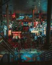 A bustling casino floor filled with groups of slot machines and people walking around. The atmosphere is vibrant with neon lights and illuminated signs. The interior has a ceiling supported by large pillars and there are stairs leading down to the gaming area.