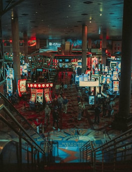 A bustling casino floor filled with groups of slot machines and people walking around. The atmosphere is vibrant with neon lights and illuminated signs. The interior has a ceiling supported by large pillars and there are stairs leading down to the gaming area.